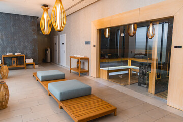 Modern spa area featuring a glass enclosed sauna, wooden benches with light blue cushions, pendant lights, and light colored tiled flooring.