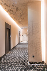 Elegant hotel hallway featuring a gray and white geometric carpet, map like ceiling artwork, recessed lighting, and dark colored doors.