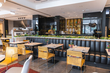 Elegant restaurant interior with yellow chairs, black banquette, floral decor, illuminated bar, open kitchen, greenery, and large windows with city views.