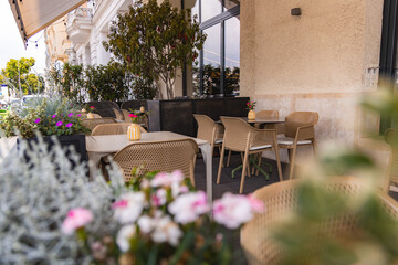 Outdoor seating area with beige wicker chairs, tables, potted plants, and flowers on a wooden deck. Beige stucco wall and glass windows frame the space.