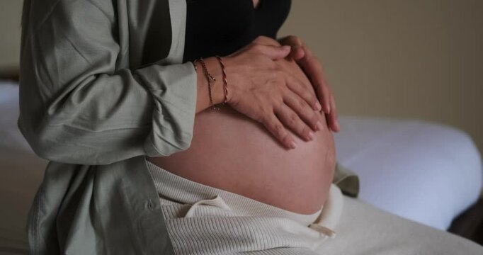 Close up of pregnant woman belly. She is sitting on bed in her bedroom and gently stroking her round belly in anticipation of the baby arrival. Third trimester of pregnancy, health care and childbirth