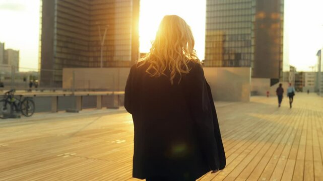 Businesswoman walks confidently toward office building, viewed from behind. Sunset light outlines her silhouette. Power, ambition, and purpose in every step — modern leadership in motion.