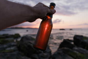 Person holding a beer bottle against the sunset light at the seaside