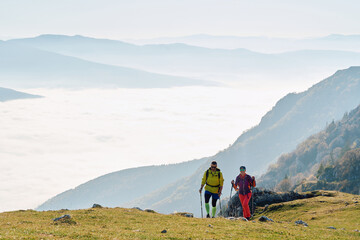Obraz premium Couple hikers trek up a rocky mountain trail, enjoying a scenic view above the clouds, with majestic peaks in the background.