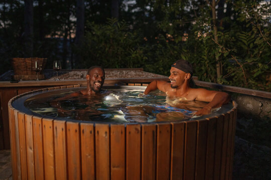 African American LGBT romantic gay couple relaxing in a private outdoor hot tub at night, enjoying glamping spa luxury drinking tasty red wine.