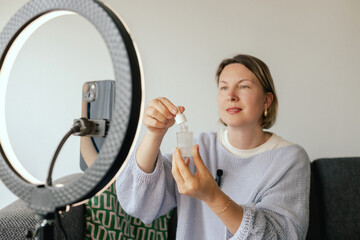 Middle-aged woman beauty blogger filming skincare product unboxing at home with ring light....
