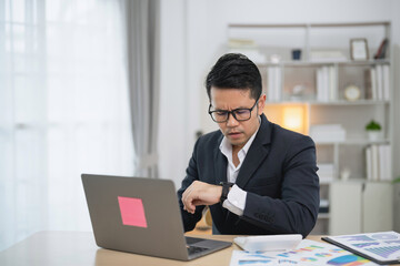 Fototapeta premium Focused businessman checking watch while working on laptop in modern office setting with documents, desk, and natural light