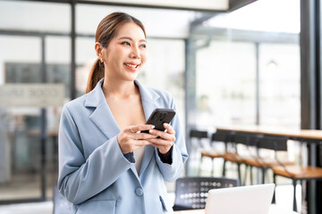 Fototapeta premium Young businesswoman standing in office using her phone and looking away outside.