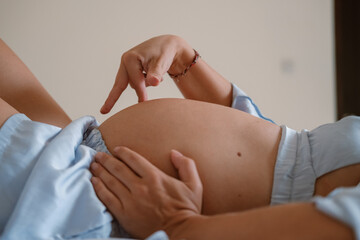 Portrait of a pregnant young woman. She lies on the bed in her pajamas and strokes her round belly in anticipation of the arrival of the baby. Third trimester of pregnancy, medical care and childbirth