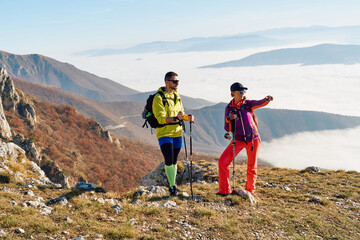 Couple hikers trek up a rocky mountain trail, enjoying a scenic view above the clouds, with majestic peaks in the background.