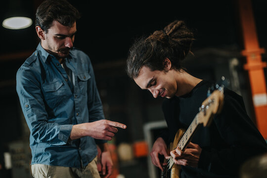 A scene showing one musician playing guitar while another gestures, providing guidance. Capture of a rehearsal atmosphere emphasizing artistry and collaboration.