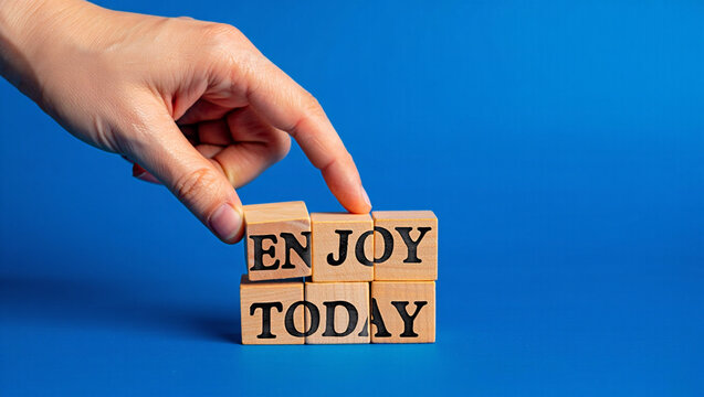 A hand places wooden blocks spelling out the words enjoy today against a blue background