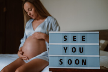 In the foreground is a sign saying see you soon, in the background is a pregnant woman in pajamas,...