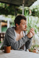 Fototapeta premium Casual scene of a man in a hoodie seated outside, lighting a cigarette, with a mug on the table and green foliage in the background.