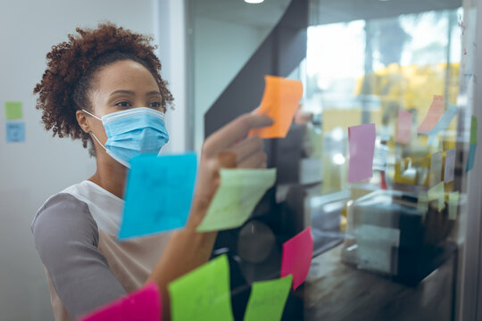 Mid-adult woman wearing blouse, medical mask placing orange note among notes on panel in office