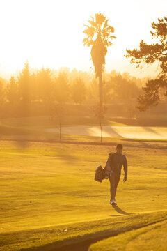 Male in twenties walking along golf course fairway during golden hour carrying golf bag with clubs