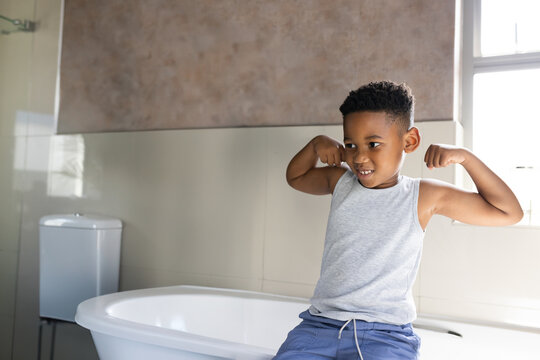 African American boy flexing arms while sitting on bathtub in home bathroom by window, copy space