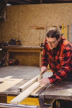 Senior male carpenter guiding plank through table saw in woodshop wearing flannel shirt, copy space