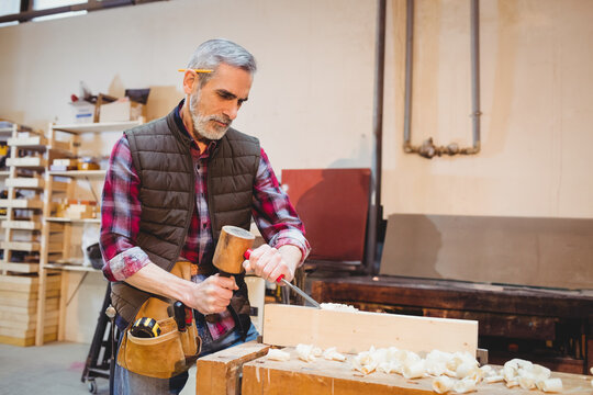 Mature male woodworker wearing plaid shirt carving block with chisel at woodworking workbench