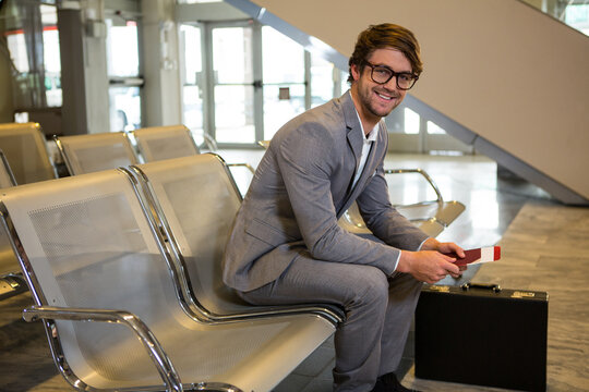 Male traveler in suit sitting at airport holding passport with briefcase by escalator, copy space