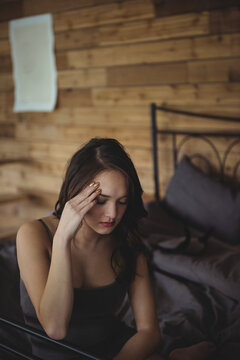 Asian woman sitting on metal bed frame pressing temple in wood-panel bedroom, copy space