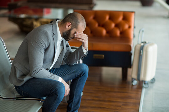 Man is sitting hunched forward on perforated metal bench in airport lounge beside rolling suitcase