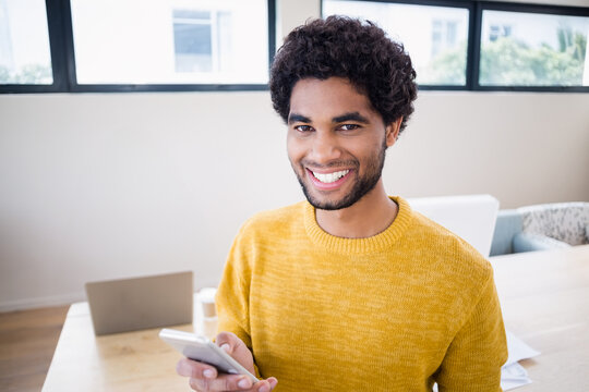 African American man smiling, holding smartphone at wooden table in office with laptop, mug, papers