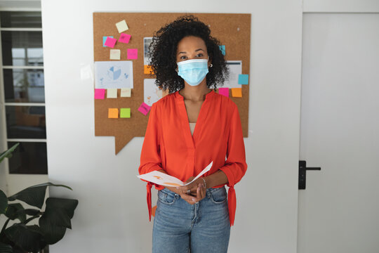 African American woman wearing medical mask reviewing notebook and charts on corkboard in office