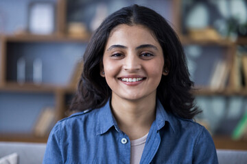 Close-up portrait of a young smiling Indian woman in a denim shirt indoors, standing and smiling at...
