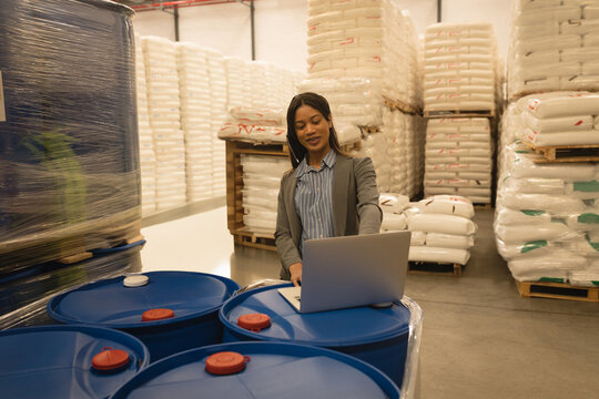 African American woman in grey blazer checking laptop at industrial drums in warehouse by pallets