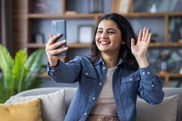 Smiling young Indian woman sitting on sofa at home, talking on video call and greeting on mobile...