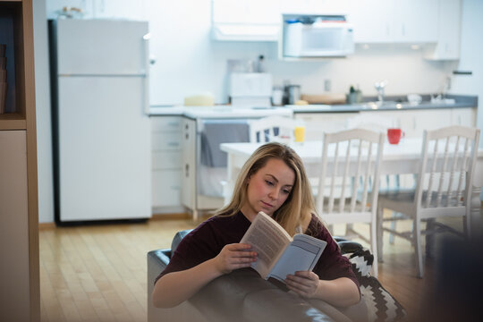 Woman reading paperback book while sitting on dark sofa in open plan living area, copy space