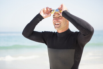 Man wearing black wetsuit adjusting yellow swim goggles while standing on sandy beach shoreline