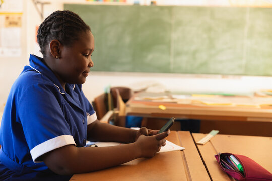 School student sitting at wooden desk using smartphone in classroom, with stationery on desk