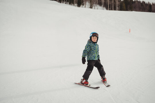 Boy descending ski slope in snowplow stance near red marker, wearing patterned jacket and helmet