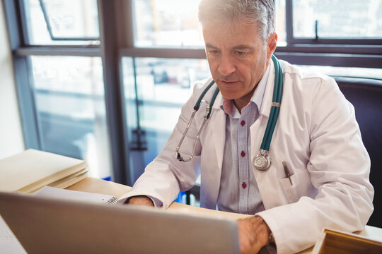 Senior male doctor typing on laptop at medical office overlooking city view with stethoscope
