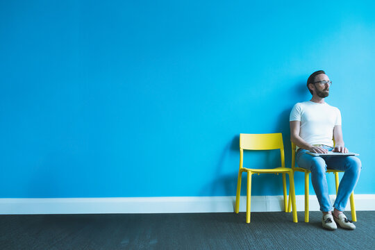 Man sitting on bright yellow chair next to empty yellow chair against blue wall, holding laptop