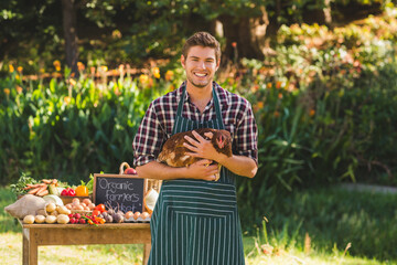 Man wearing striped apron cradling brown chicken at farm stand with produce, chalkboard sign