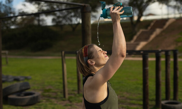 Female athlete lifting water bottle, pouring water over face in fitness park by pull-up bars, tires