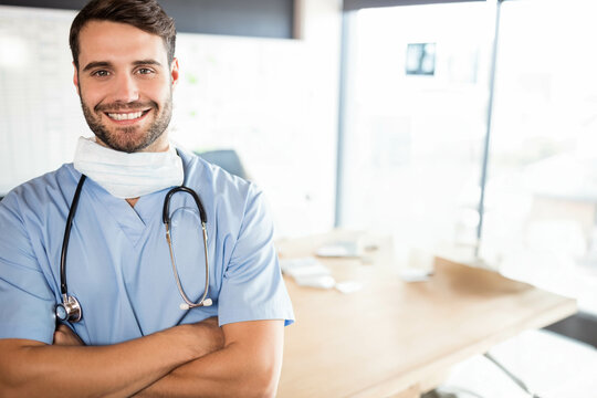 Draping stethoscope and hanging surgical mask, doctor standing with arms crossed by desk in clinic