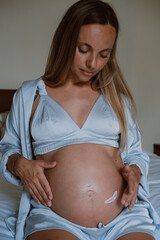 Close up of pregnant woman big belly, she is sitting on bed in pajamas and applying moisturizing...