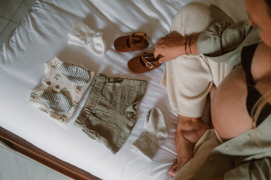 Portrait of pregnant woman with big belly, who is sitting on bed and putting away baby clothes, getting ready for maternity hospital. She examines each thing and strokes belly, waiting baby to appear.