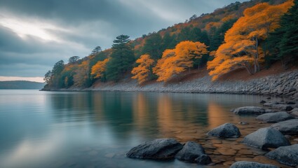 Obraz premium A tranquil lakeside scene featuring a rocky shoreline and trees. The still water mirrors the cloudy sky, with foliage adorning the hillside, producing a peaceful ambiance.