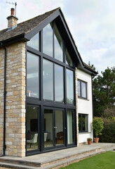 A striking modern house extension featuring a two-story glass wall with black frames, blending rustic sandstone cladding with a clean white render for a light-filled home.
