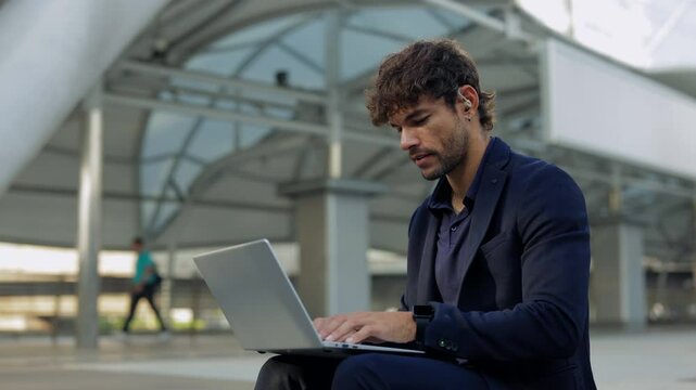 Portrait of young Hispanic man with hearing aid works on laptop at office building. Latin businessman with disability writes report on notebook computer sitting on downtown street
