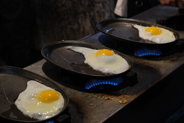 Three sunny side up eggs being cooked on individual hot plates over open flames. 