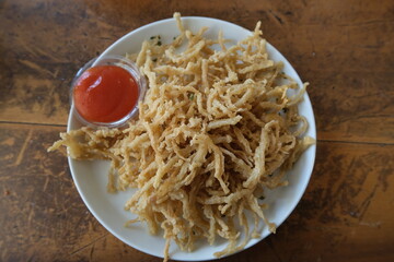 Crispy fried enoki mushrooms served on a white plate with red chili sauce on rustic wooden table.