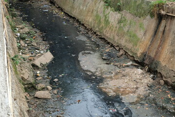 Polluted urban drainage canal filled with black water, plastic waste, and sediment buildup,...