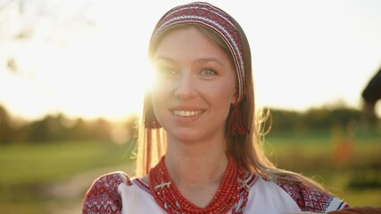 Close-up portrait of a young woman in traditional red and white folk clothing, smiling warmly while standing outdoors near a village house in golden evening light. - Powered by Adobe