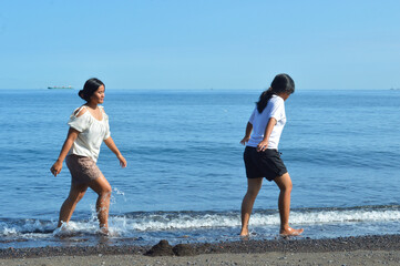 Two teenage girls play in the shallow waves at the beach, dipping their toes into the refreshing seawater while walking along the shoreline, enjoying the peaceful morning.